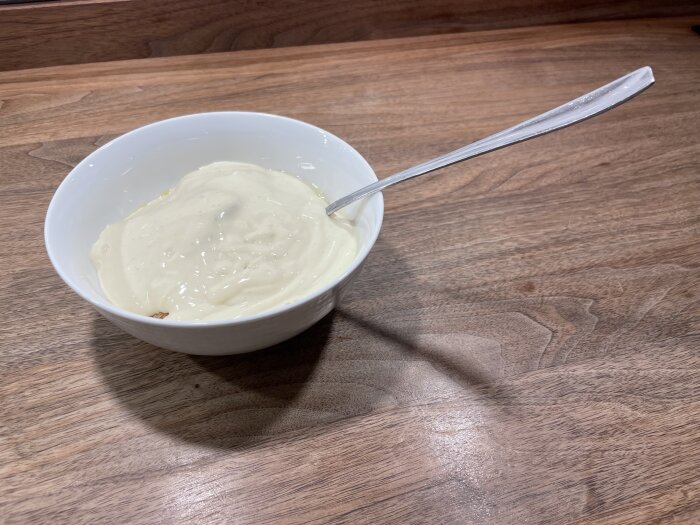 En vit skål med krämig vaniljyoghurt och en sked, på ett brunt träbord.