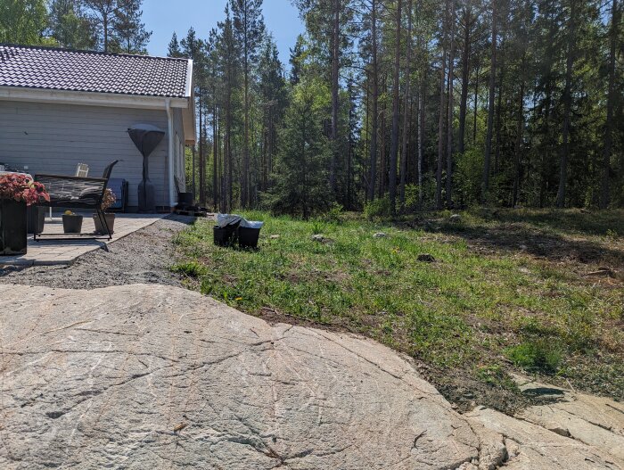 Ett hus med trädäck, grill, skog i bakgrunden, sten i förgrunden, soligt väder.