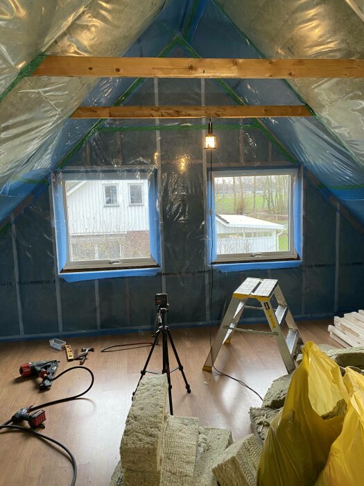 Renovering pågår, isolering, stege, tripod, verktyg, plasttäckt tak och väggar, oavslutat byggprojekt inomhus.