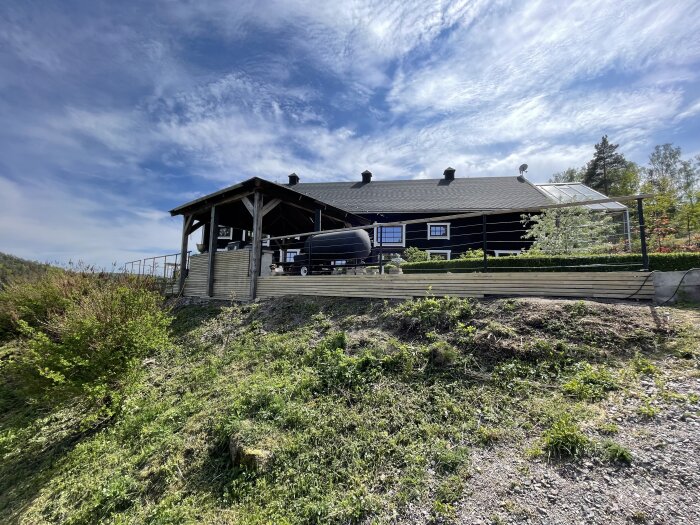 En stor trästuga med veranda, grill, och skogsbakgrund under en blå himmel med vita moln.