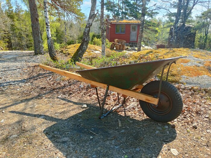 Skottkärra framför lantligt rött hus, skogsmiljö, soligt, mossiga stenar, vedtrave, tallar och björkar.