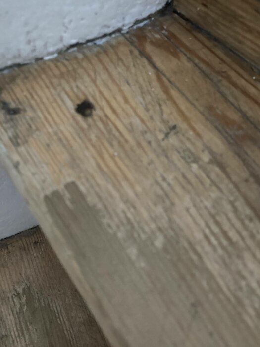 Närbild på ett trägolv och en vägg med spikhål och nötta kanter.