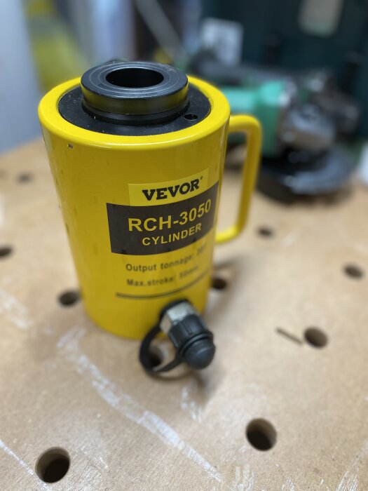 Gul hydraulcylinder av märket VEVOR, modell RCH-3050, på en arbetsbänk, grön maskin i bakgrunden, suddig.