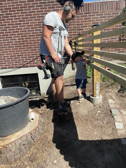 Man och litet barn bygger staket tillsammans utomhus vid en tegelvägg på en solig dag.