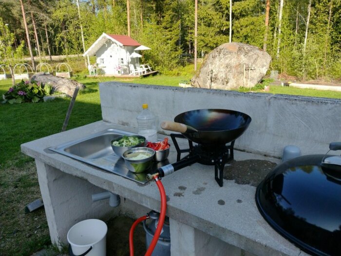 Utomhuskök med wokpanna på gasolbrännare, grönsaker redo, lantlig trädgård i bakgrunden.