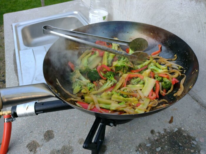 Wokpanna med grönsaker på gasbrännare utomhus. Matlagning, ånga, färgglatt, broccoli, paprika, zucchini, matstekning.