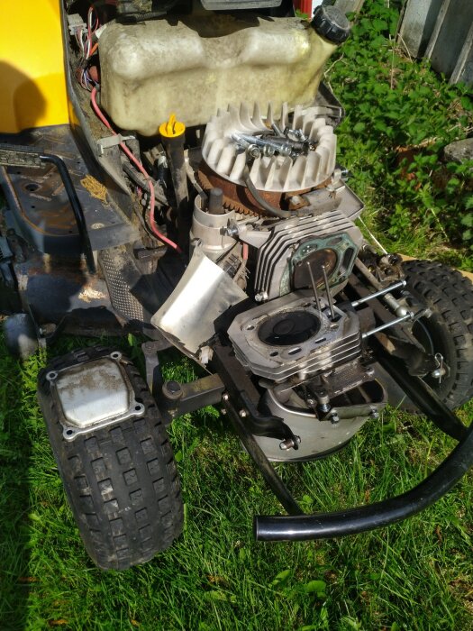 Delvis demonterad motor på gräsmaskin, utsatt för ljus, grönt gräs i bakgrunden.