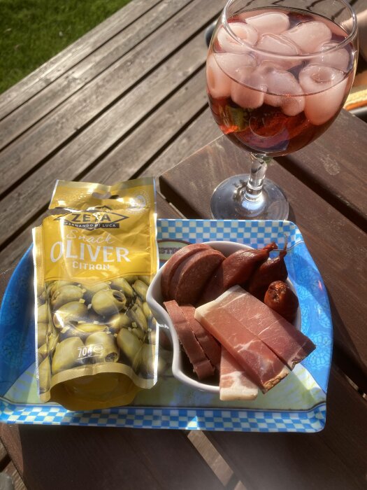 Ett glas med en roséfärgad dryck och isbitar, förpackade oliver och en blandning av kött på ett fat utomhus.