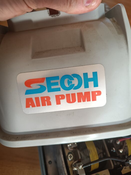 En person håller i en luftpump märkt "SECOH AIR PUMP," med synliga elkomponenter under.