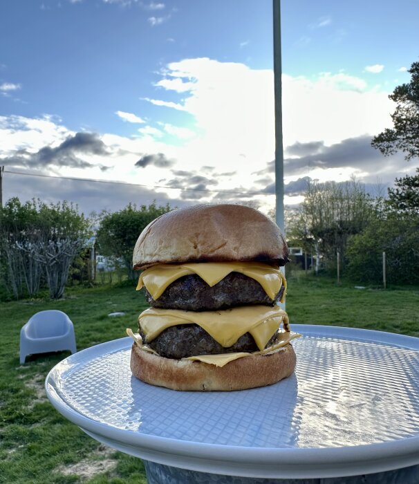 En stor cheeseburgare utomhus på ett bord, med himmel och trädgård i bakgrunden.