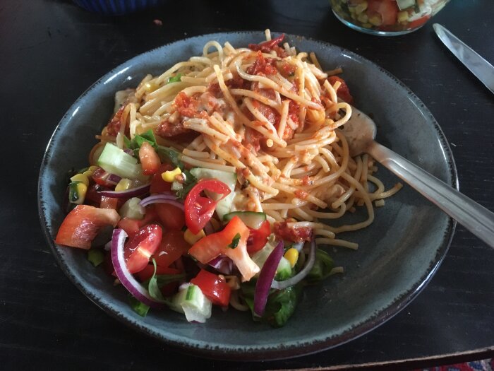 Tallrik med spaghetti, tomatsås, färgglad sallad, gaffel, mörk bordsyta.