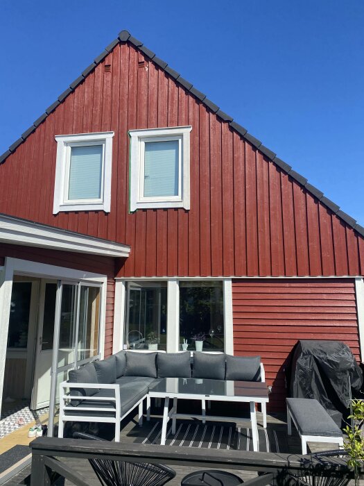 Röd trähusfasad, vita fönster, uteplats med loungegrupp, klarblå himmel, grillöverdrag.