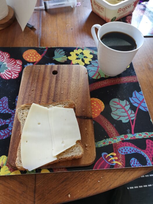 Brödskiva med ost och kaffekopp på mönstrad duk och träskärbräda. Bakgrund med smörask. Enkel frukost eller fika.