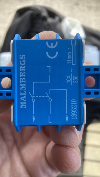 Blå elektrisk komponent med skruvanslutningar och elektriskt schema, hålls av en hand, CE-märkt, MALMBERGS-märke.