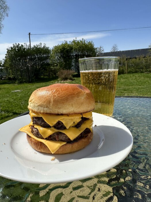 Dubbel cheeseburgare på vit tallrik och ett glas läsk utomhus på solig dag.