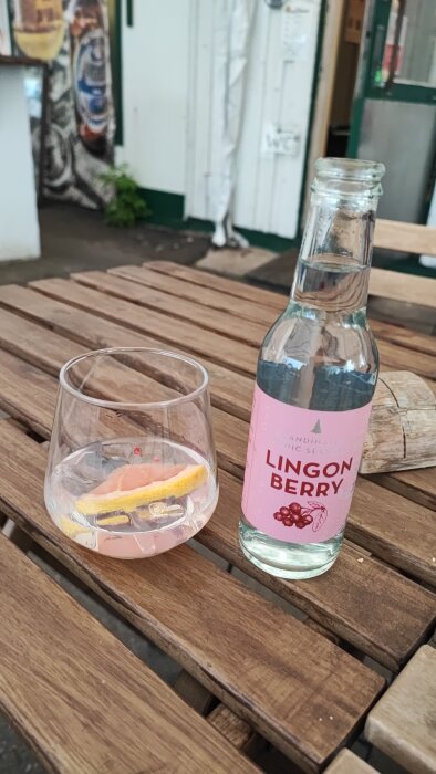 En tom lingonberry tonic-flaska bredvid ett glas med tonic och citronskiva på ett träbord utomhus.