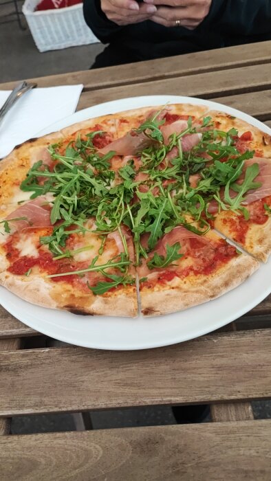 Pizza med skinka och ruccola på ett vitt fat, person med korsade händer i bakgrunden.