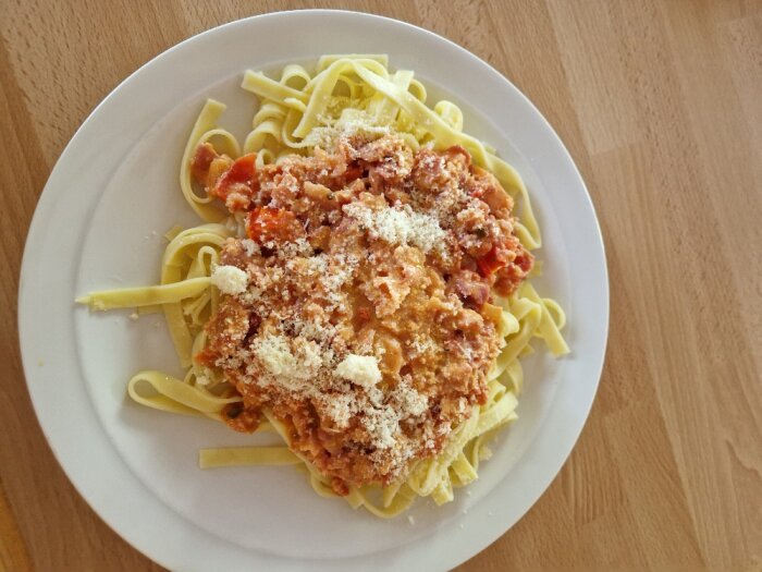 Tallrik med pasta, tomatsås, köttfärs, strödd parmesan, träbord i bakgrunden.