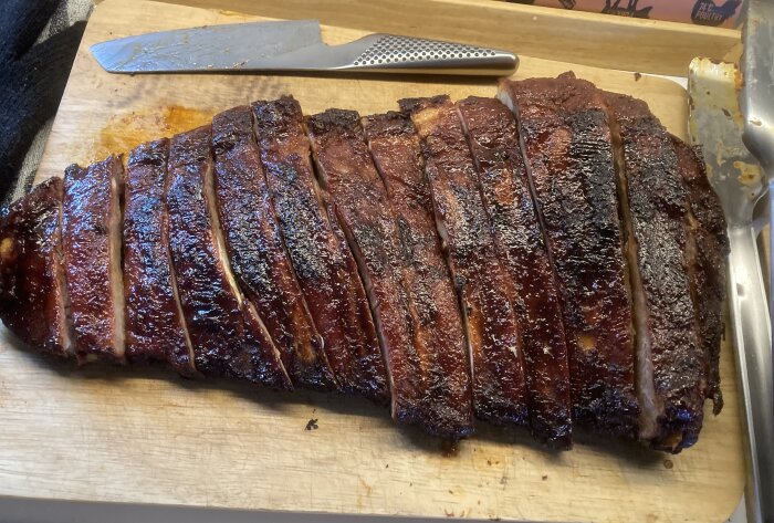 Grillade revbensspjäll, skurna och klara på träskärbräda med knivar. Glansigt, brun-svart yta som antyder karamellisering.