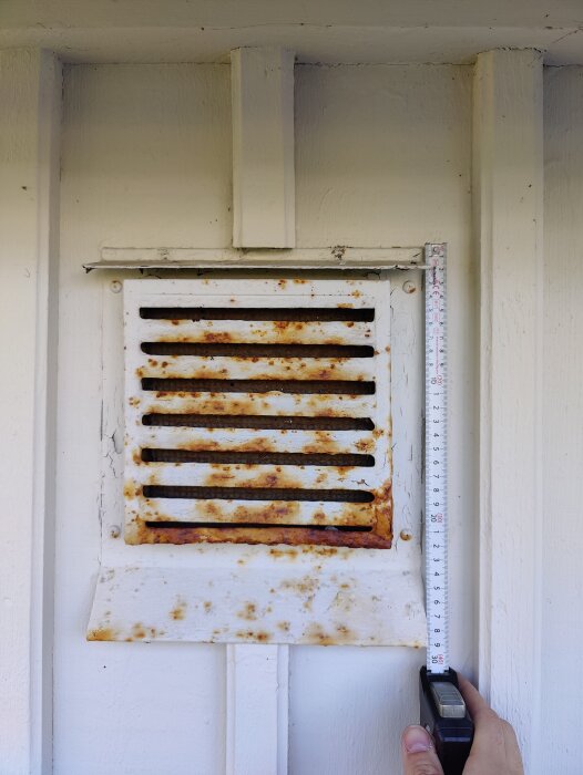 En rostig ventilationsgaller på en vit vägg mäts med en tumstock.