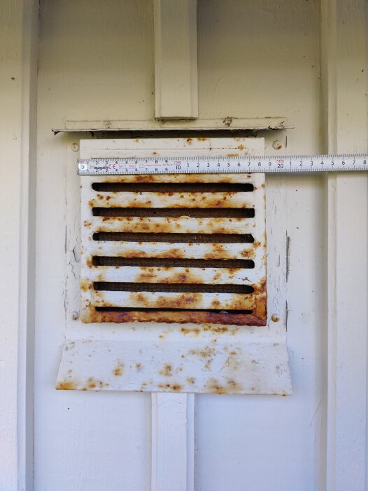 En rostig vit ventilationsgaller på en vägg med en mätsticka framför.