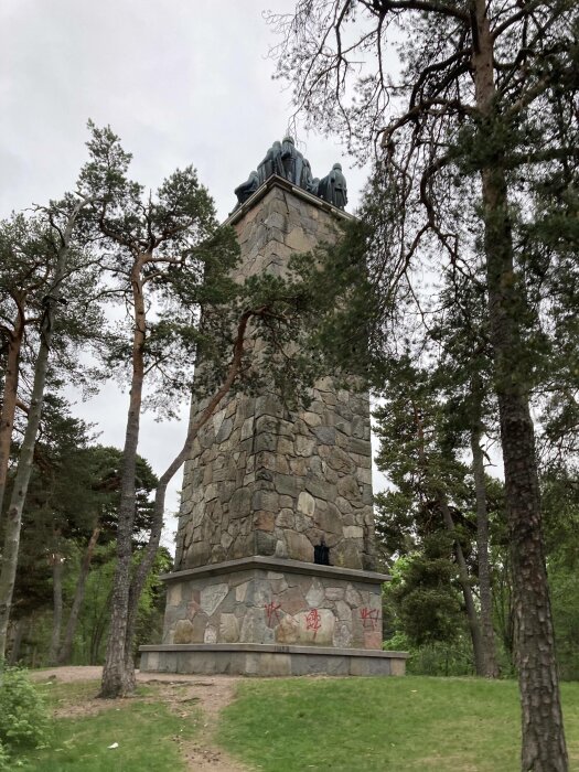 Stentorn i skog med statyer på toppen, grå himmel, graffiti på basen, gröna träd runtomkring.