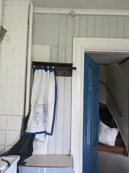 Äldre interiör, skadad vägg, hängande handduk, blå dörr, trätrappa, hemmiljö, ljusinfall, slitet.