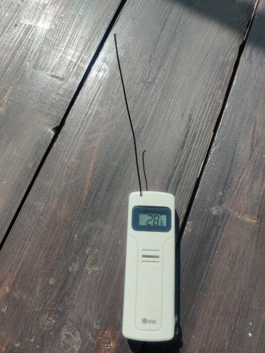 Digital termometer visar 28.1 grader Celsius på en träyta under solljus.