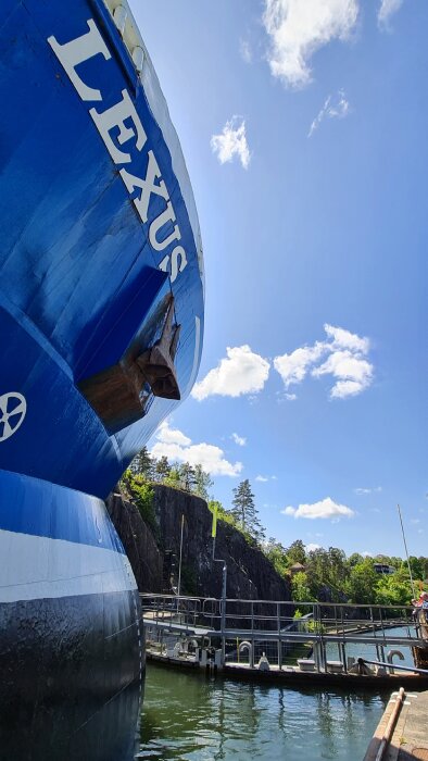 Stort blått fartyg med namn "LEXUS", förtöjt vid klippig hamn, klar himmel, del av brygga synlig.