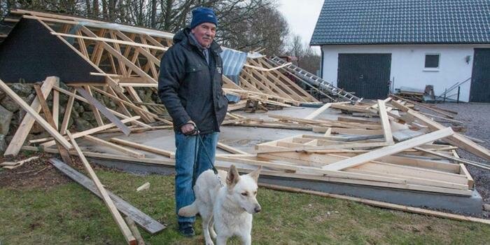 Man med hund står framför kollapsad träkonstruktion, troligen en byggnad, bland bråte och spillror.