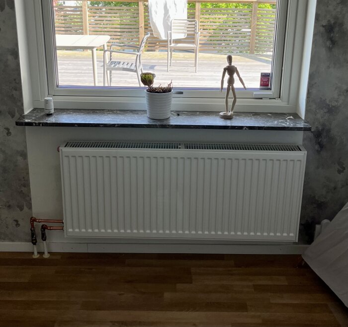 Ett fönster med stenfönsterbräda, en vit kruka med växt, en träfigur, en radiator, och en terrass utanför.