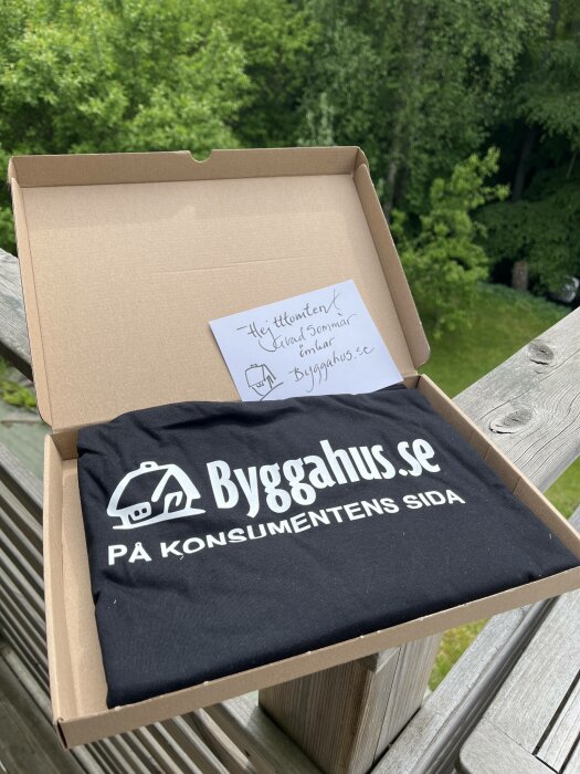 T-shirt i kartong med logotyp "Byggahus.se På konsumentens sida", handskriven lapp, utomhusmiljö.