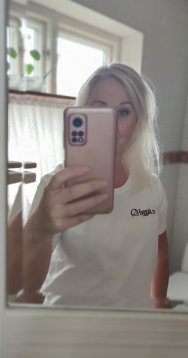 Kvinna tar selfie med mobil i spegel, dagsljus, inomhus, suddig bakgrund, vit T-shirt med logotyp.