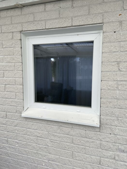 Vitt fönster i tegelvägg, reflekterande glas, skymtar mörkt interiör, inget synligt liv.