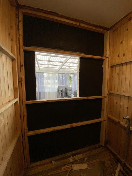 Ett fönster i en träklädd vägg med utsikt mot ett inglasat uterum. Inomhusmiljö under renovering eller ombyggnad.