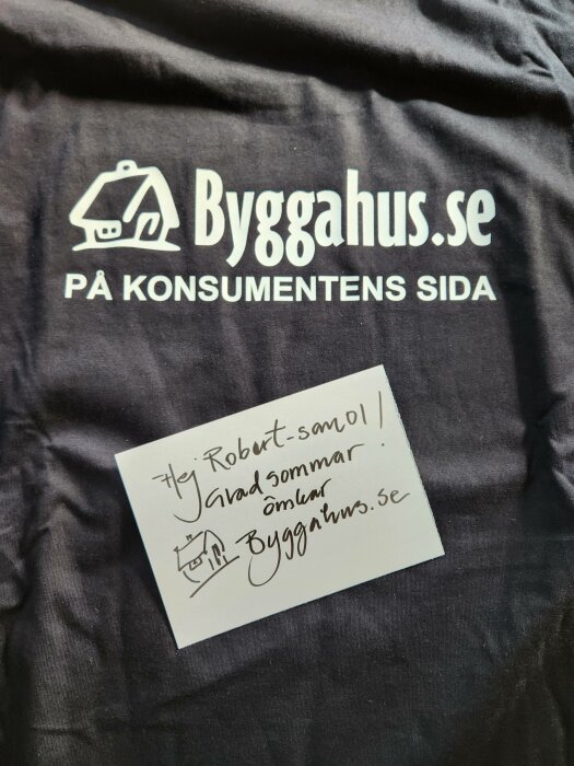 Svart t-shirt med "Bygghus.se" logotyp, handskriven hälsning på lapp, "Glad sommar" önskan, tygets textur syns.