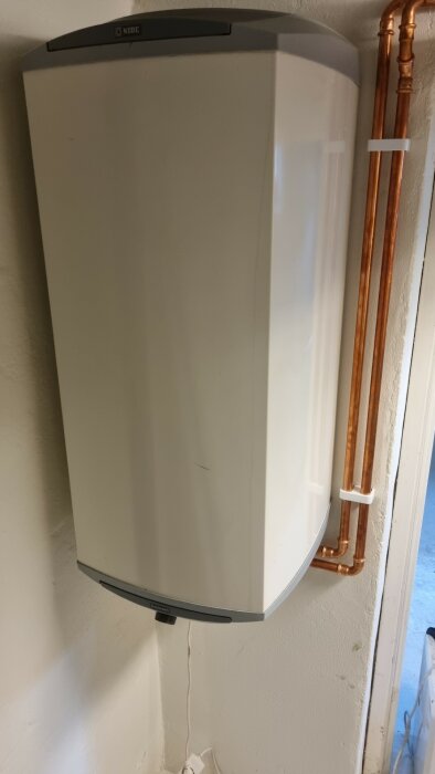 Väggmonterad varmvattenberedare med kopparledningar i ett hörn av ett rum.