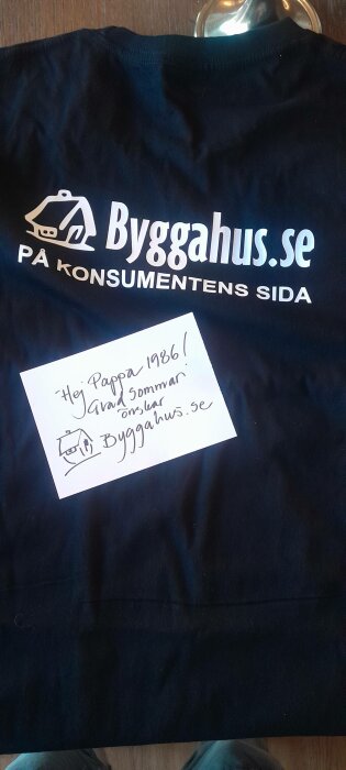 Marinblå T-shirt med "Byggahus.se" logotyp och en handskriven lapp där det står "Hej Pappa 1986! Glad sommar önskar Byggahus.se".