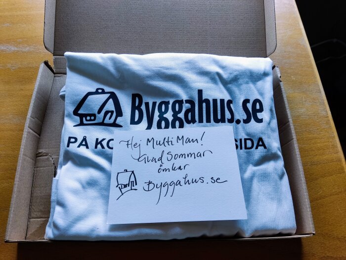 T-shirt i kartong, tryck med text och symbol, handskriven hälsning på kort, "Hej MultiMan!" "Glad sommar önskar byggaahus.se".