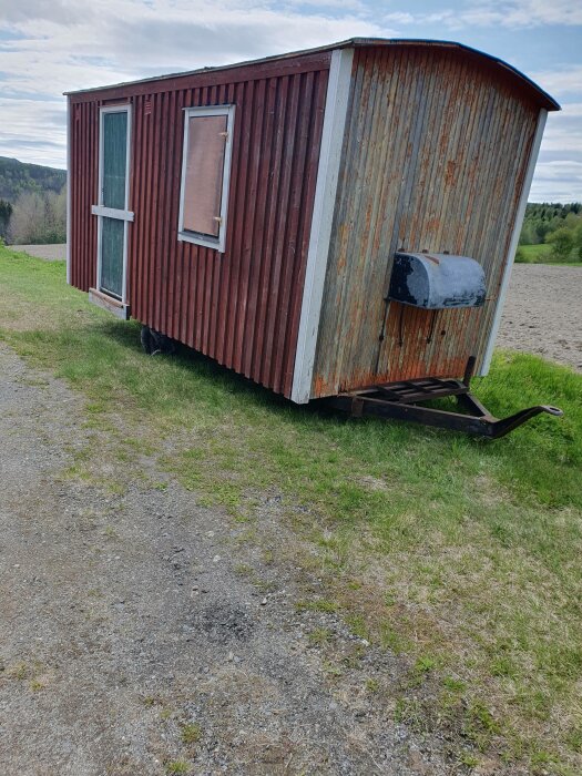 Gammal släpvagn ombyggd till bostad med rost och patina, stående i landsbygdsmiljö.