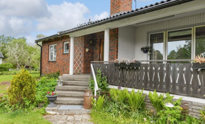 Ett tegelhus med veranda, grönska, trädgård och en delvis molnig himmel.