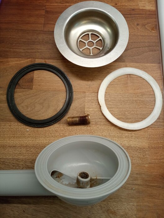 Demonterad avloppssil med packningar, skruv och monteringsdelar på ett träbord.