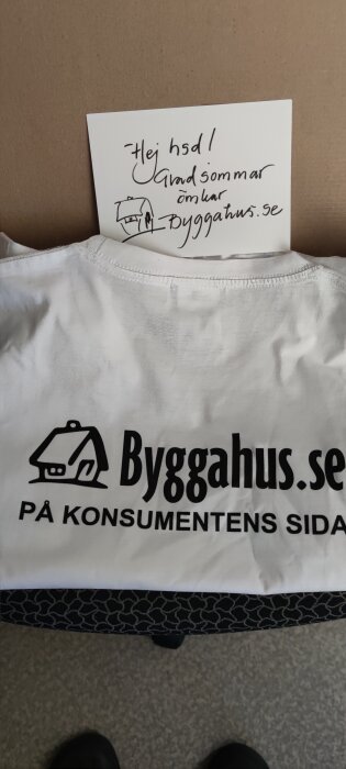 Vit t-shirt, byggnadsritning, text "Byggahus.se på konsumentens sida", handskriven hälsning, skosnok i förgrunden.