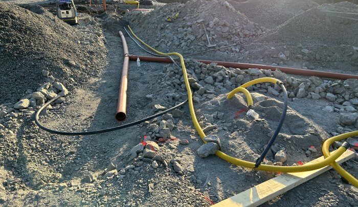 Byggarbetsplats med rör, kablar och grus. Maskiner i bakgrunden. Byggaktivitet eller infrastrukturuppgradering under solnedgång.