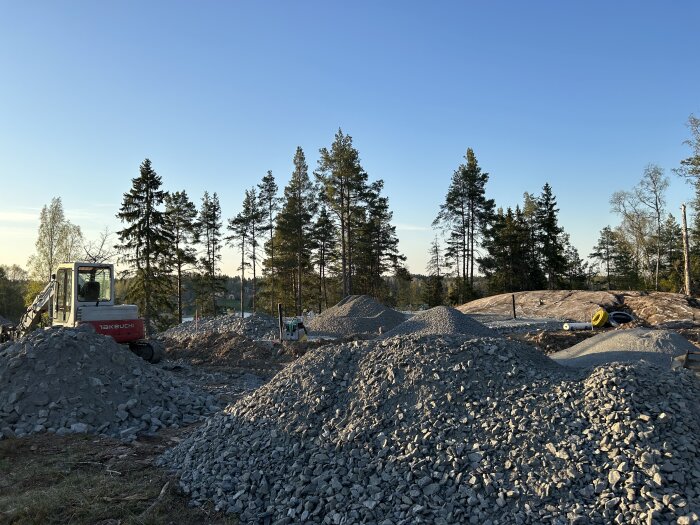 Byggarbetsplats med grävmaskin och grushögar, klar himmel, skog i bakgrunden, byggmaterial på marken.