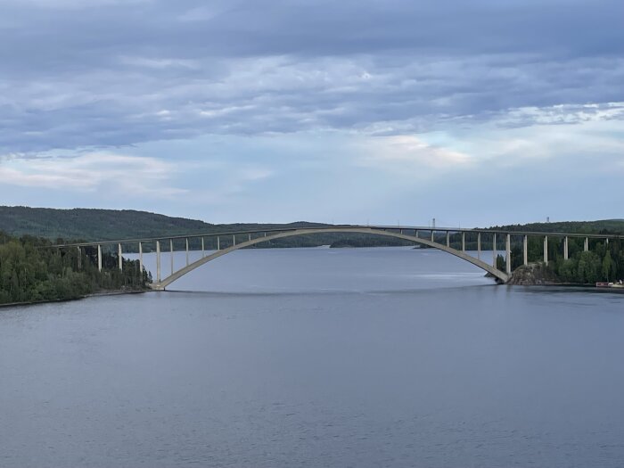 Bågbro över vatten, skogklädda kullar, molnigt himmel, reflektion på vattenytan, ingen synlig trafik.