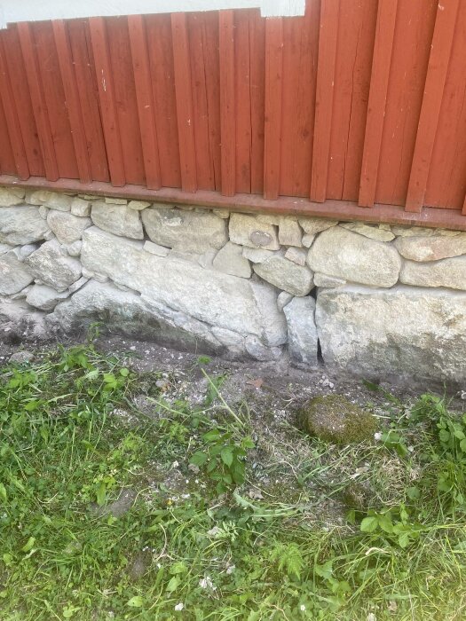 Rödmålad trävägg, stenfot, gräs och viss vegetation i förgrunden.