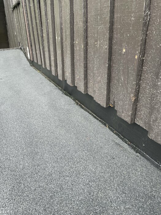 Ett brunstaket längs en asfalterad yta, skuggor synliga, slitet, utomhus under dagtid.