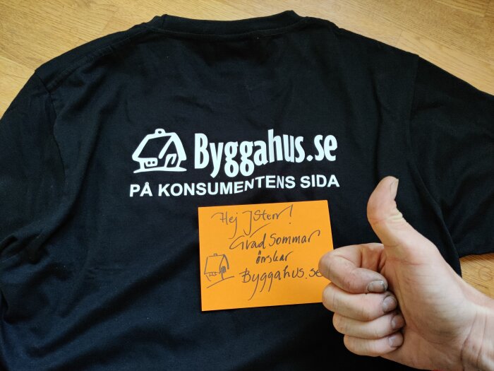 Svart T-shirt med logotyp, text, hand som håller ett orange kort med hälsning.