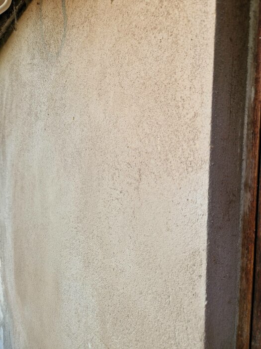 En vägg med grov puts och träram syns. Skuggor och naturligt ljus. Strukturerad yta, beige färg.